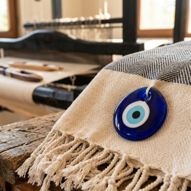 Blaues Augenamulett auf einem cremefarbenen und dunkelblauen Fransentuch, mit einem Webstuhl im Hintergrund.