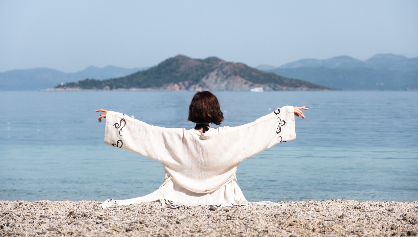 A woman wearing a natural linen kimono sits by the sea with arms wide open, symbolizing freedom, balance, and the holistic energy of fabrics.