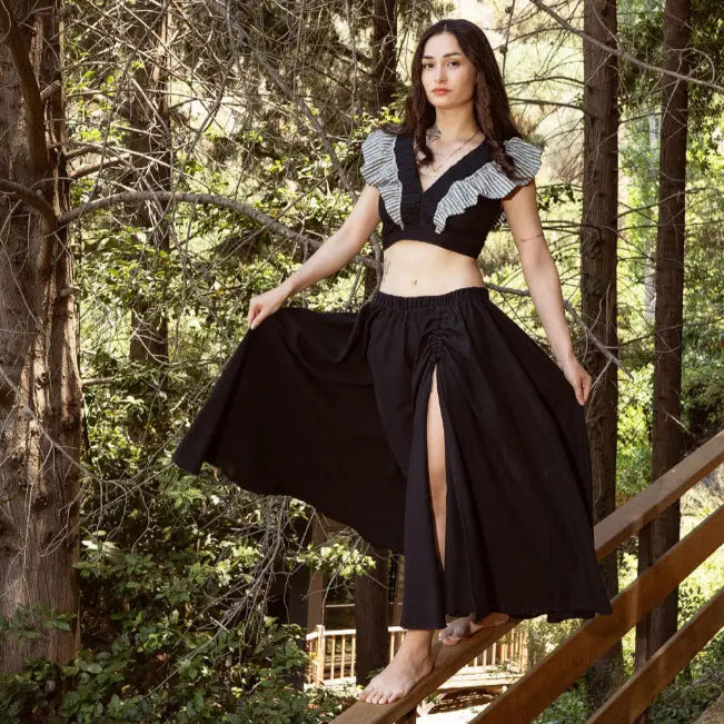 Vrouw in een zwarte crop top met patroon en een zwierige zwarte rok op een houten trap tussen bomen.