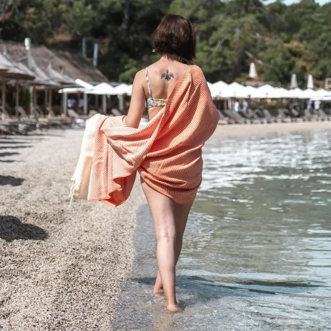 Vrouw van achteren gezien, gehuld in een koraalkleurige handdoek, lopend langs een kiezelstrand.