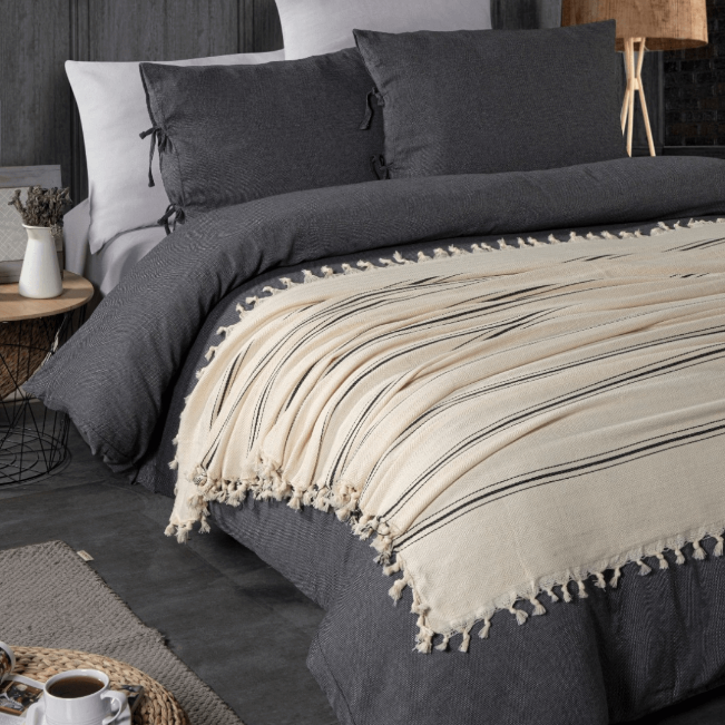 Crèmekleurige plaid met geknoopte franjes, uitgespreid over donkergrijs beddengoed.