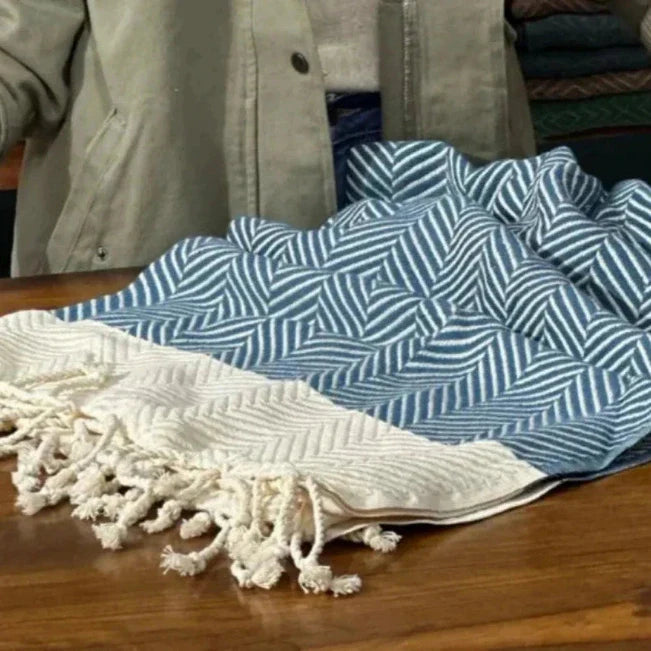 Blauw-crèmekleurige handdoek met visgraatpatroon en geknoopte franjes, uitgespreid op een houten tafel.