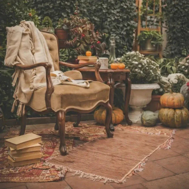 Beige plaid over een fauteuil, omringd door boeken, potplanten en pompoenen op een betegeld terras.