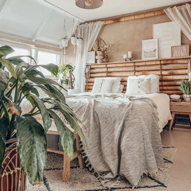 Grijze sprei met franjes op een bed met een lattenhoofdbord, lichte kussens en groene planten ernaast.