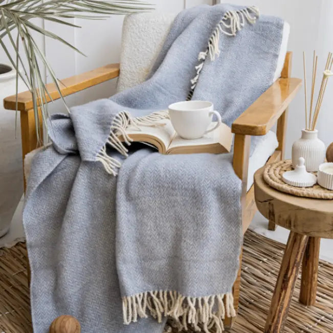 Blauwgrijze plaid met franjes op een houten fauteuil met een open boek en een witte kop.
