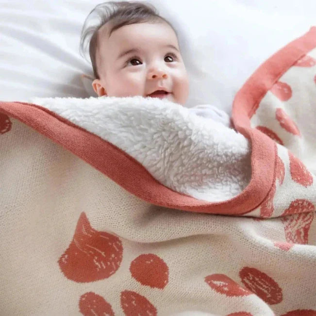 Baby kijkt omhoog onder een crèmekleurige deken met koraalkleurige patronen en een pluizige witte voering.