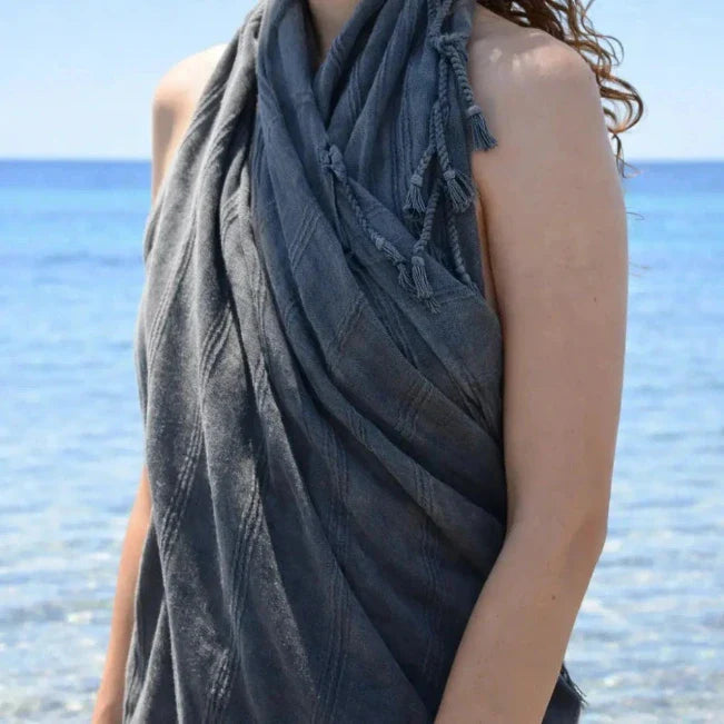 Detail van een donkerblauwe handdoek met franjes over de schouder van een vrouw aan zee.