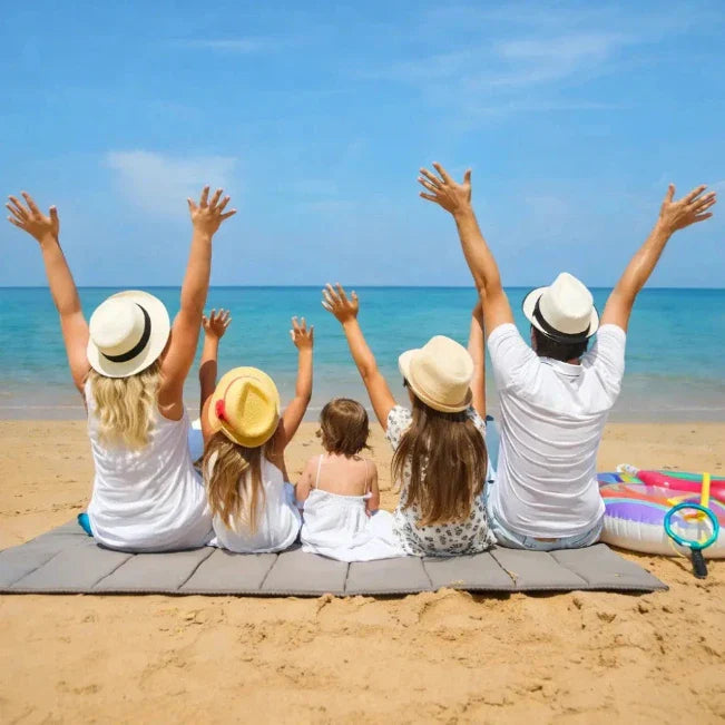 Zwei Erwachsene und drei Kinder sitzen mit erhobenen Armen am Strand und blicken aufs Meer.
