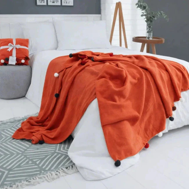 Orange Decke mit dunklen Pompons auf einem weißen Bett.