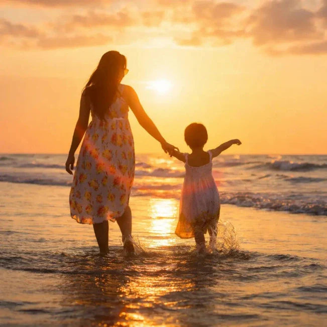 Vrouw en kind houden elkaars hand vast in ondiep zeewater bij zonsondergang.