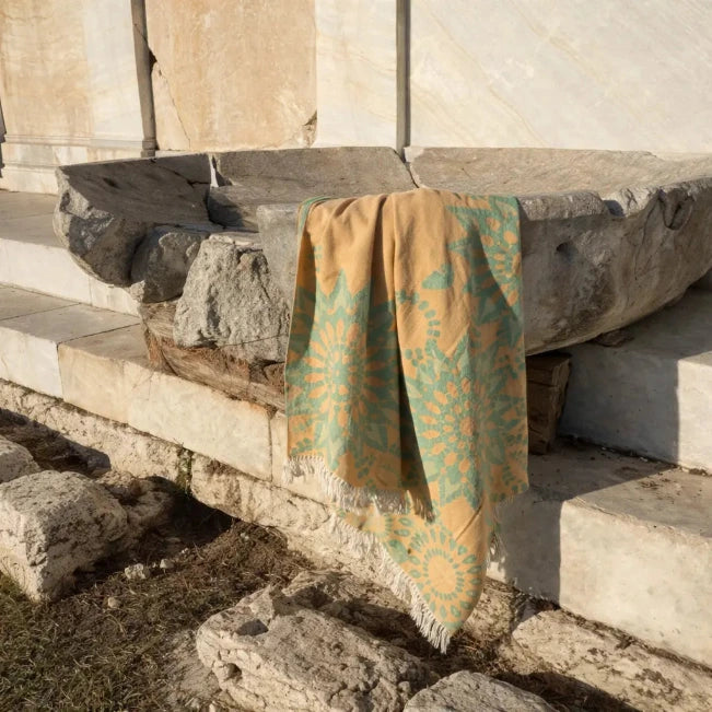 Goud-groene handdoek met dessin en franjes over verweerde stenen blokken.