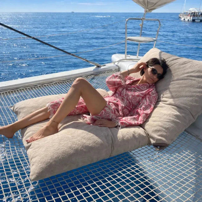 Vrouw in een roze-witte badjas met dessin ligt op kussens op een boot.