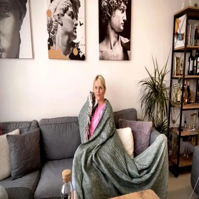 Vrouw gehuld in een groene plaid met patroon op een grijze bank, onder zwart-witte kunstprenten.