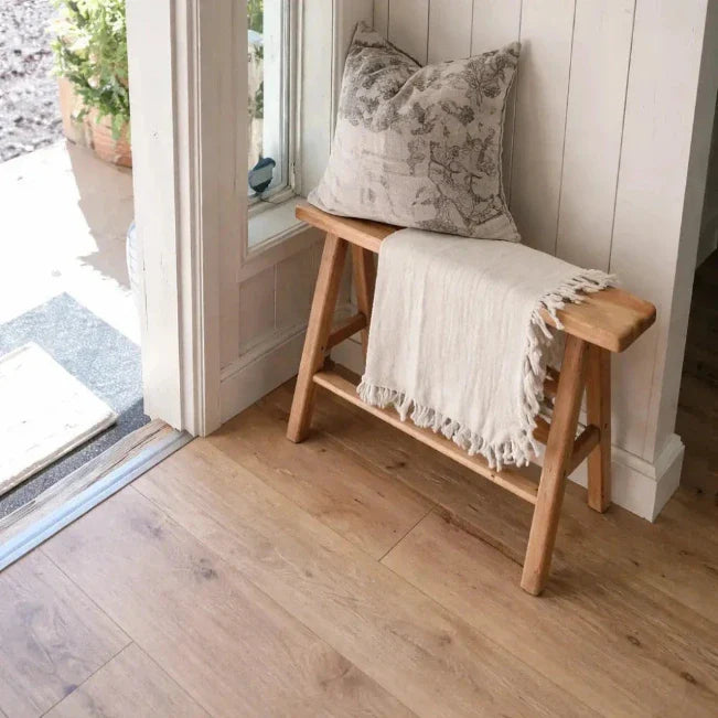 Lichte plaid met franjes en kussen met dessin op een houten bankje naast een deuropening.