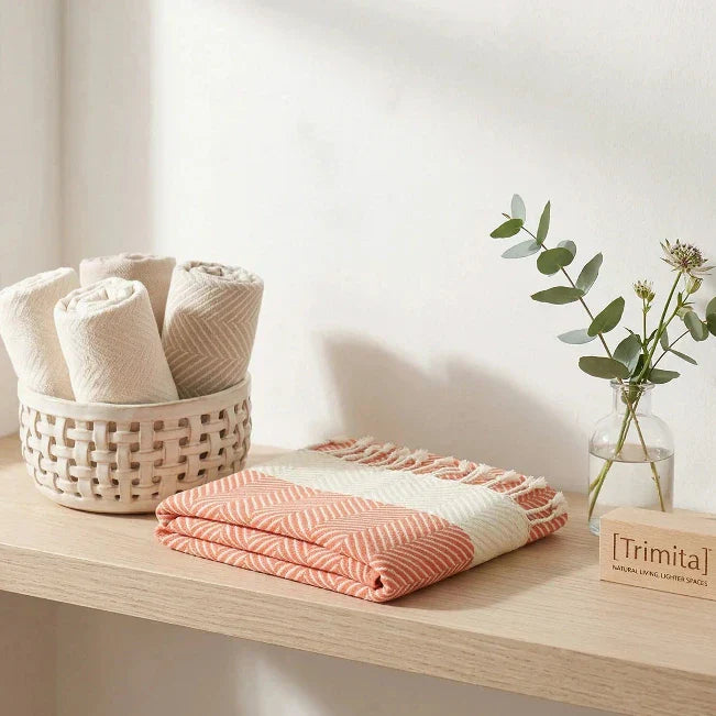Folded Trimita Turkish towel with orange and white herringbone pattern displayed on a wooden shelf next to eucalyptus leaves in a minimalist bathroom setting.