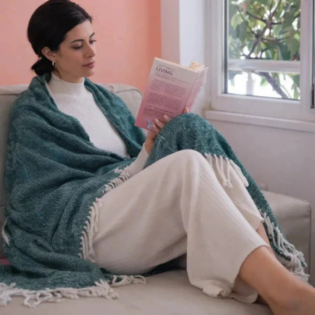 Vrouw leest naast een raam, gewikkeld in een blauwgroene plaid met franjes.