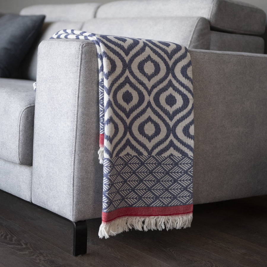 Plaid bleu marine et crème à motifs géométriques, bordure rouge et franges blanches sur un fauteuil gris.