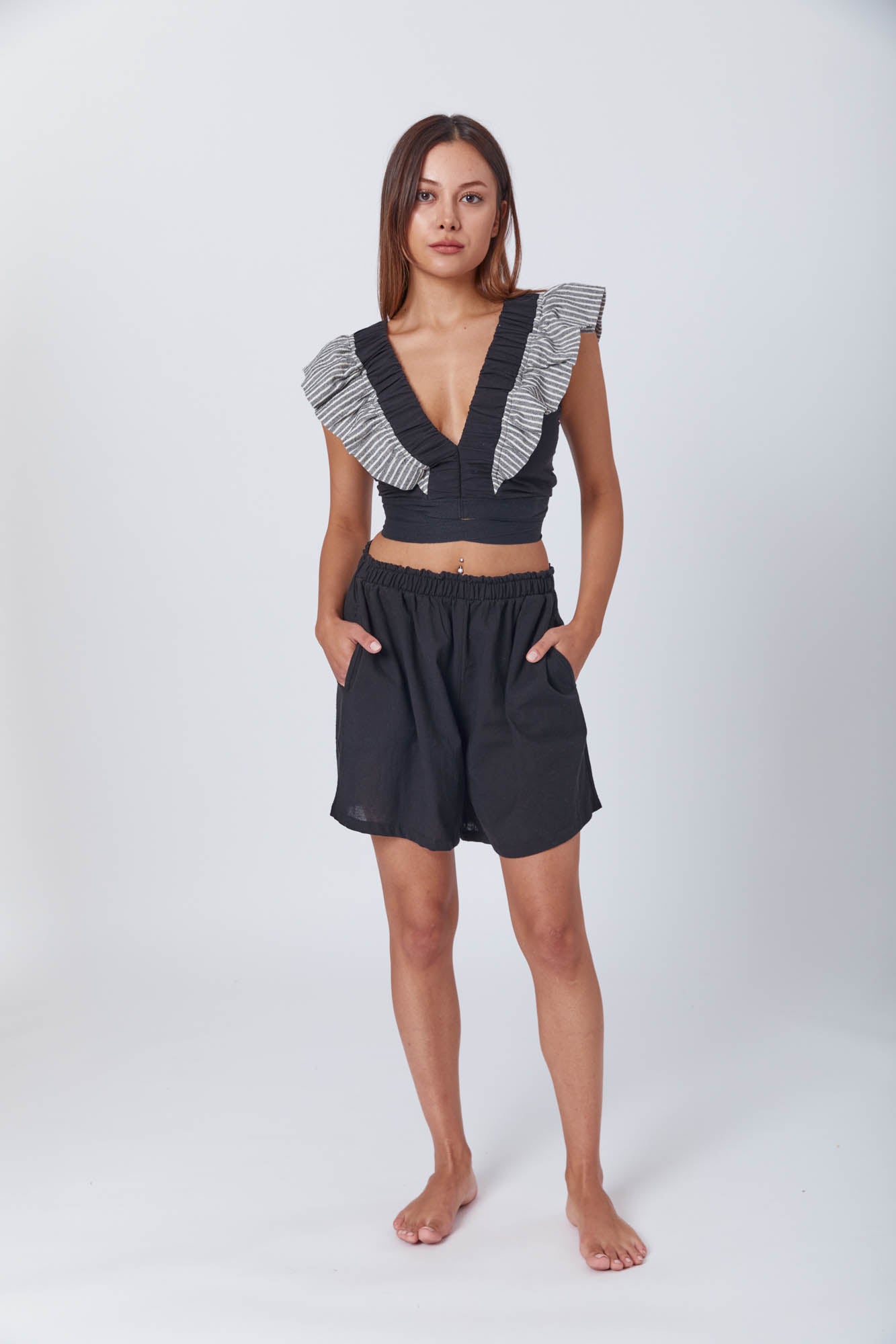 Model wearing a black sleeveless crop top with ruffled striped shoulder details and deep V-neck, paired with high-waisted black pants