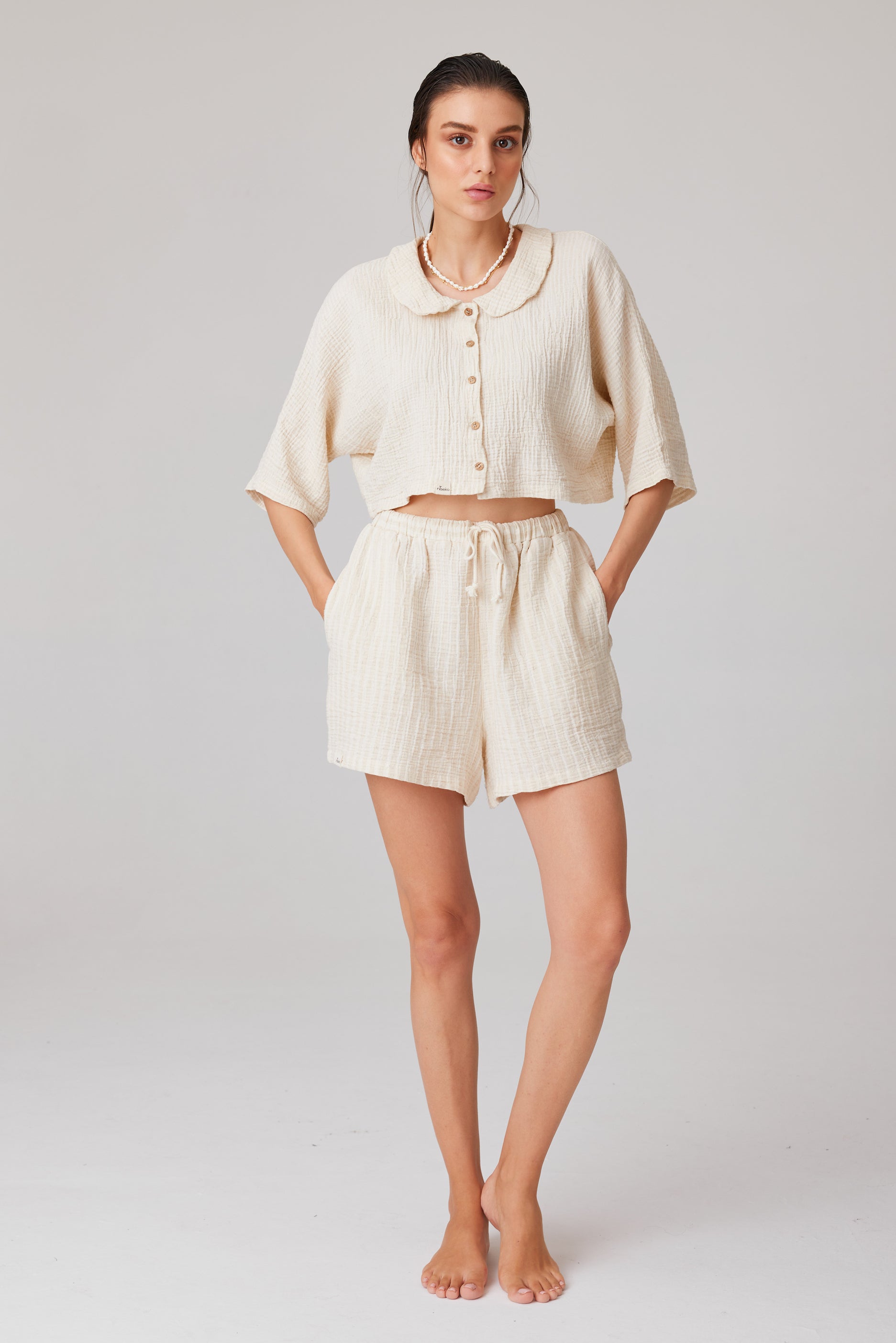 Young woman dressed in a soft-textured, off-white cotton set with a short-sleeved collared crop top and flowy shorts with pockets, standing barefoot against a light background