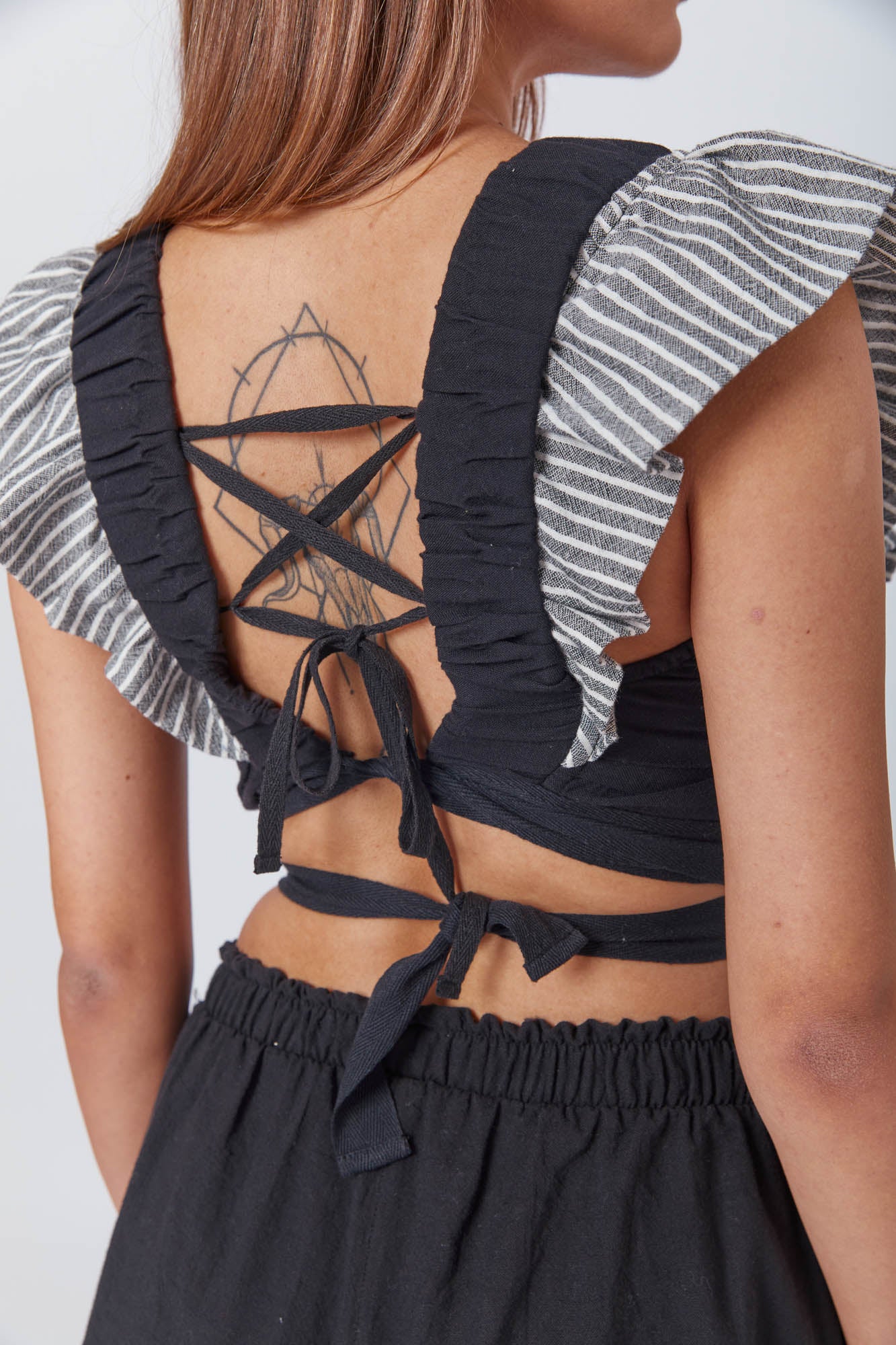 Person with straight brown hair wearing a fitted black top and striped ruffle sleeves, and casual black trousers