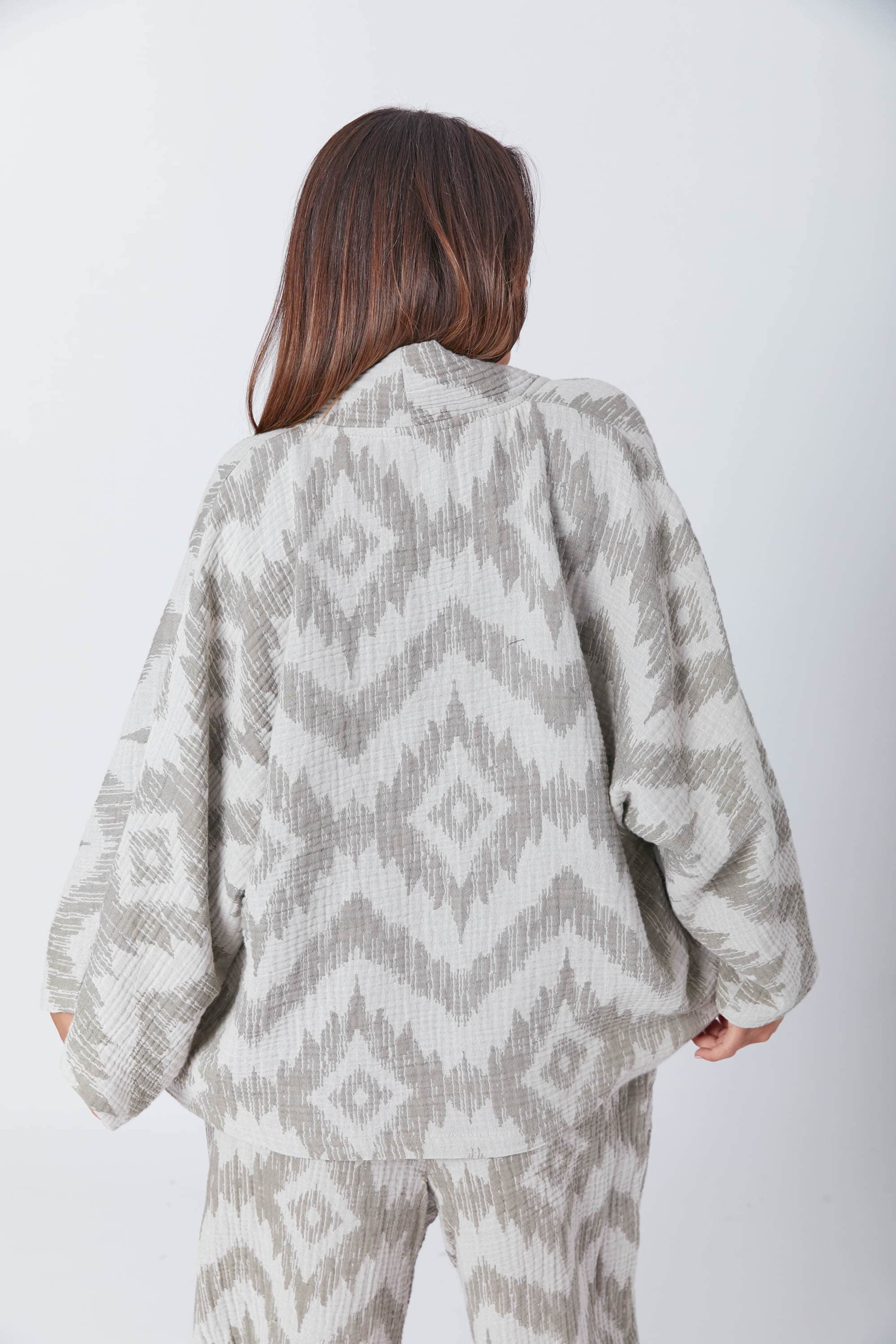 Back view of a woman wearing a draped and textured Guru Green muslin jacket decorated with ikat patterns. With its wide sleeves and draped fit, this jacket can be worn comfortably over matching trousers.