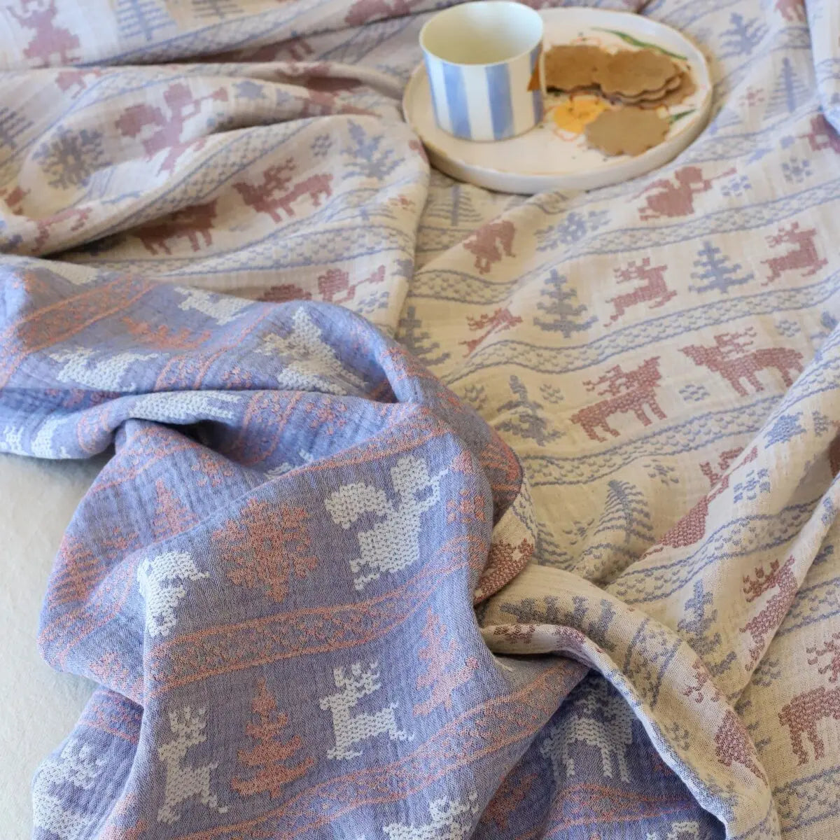 Patterned bedpread with reindeer design draped over a surface, with a cup and plate in the background.