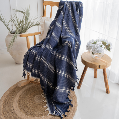 Trimita Yasmin Bedspread Navy striped blanket draped over a wooden chair in a bright room with a plant and small table.
