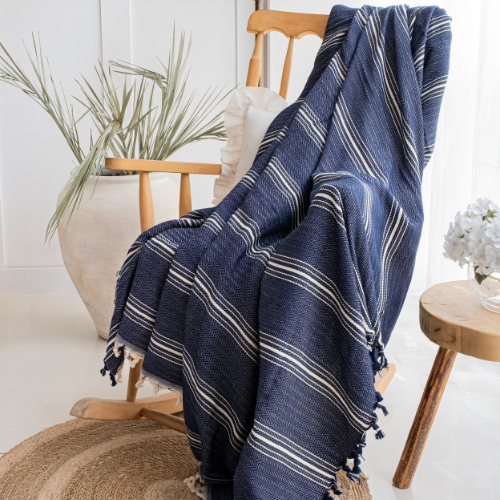 Trimita Yasmin Bedspread Navy blue striped blanket draped over a wooden chair in a bright room with plants and a small table.