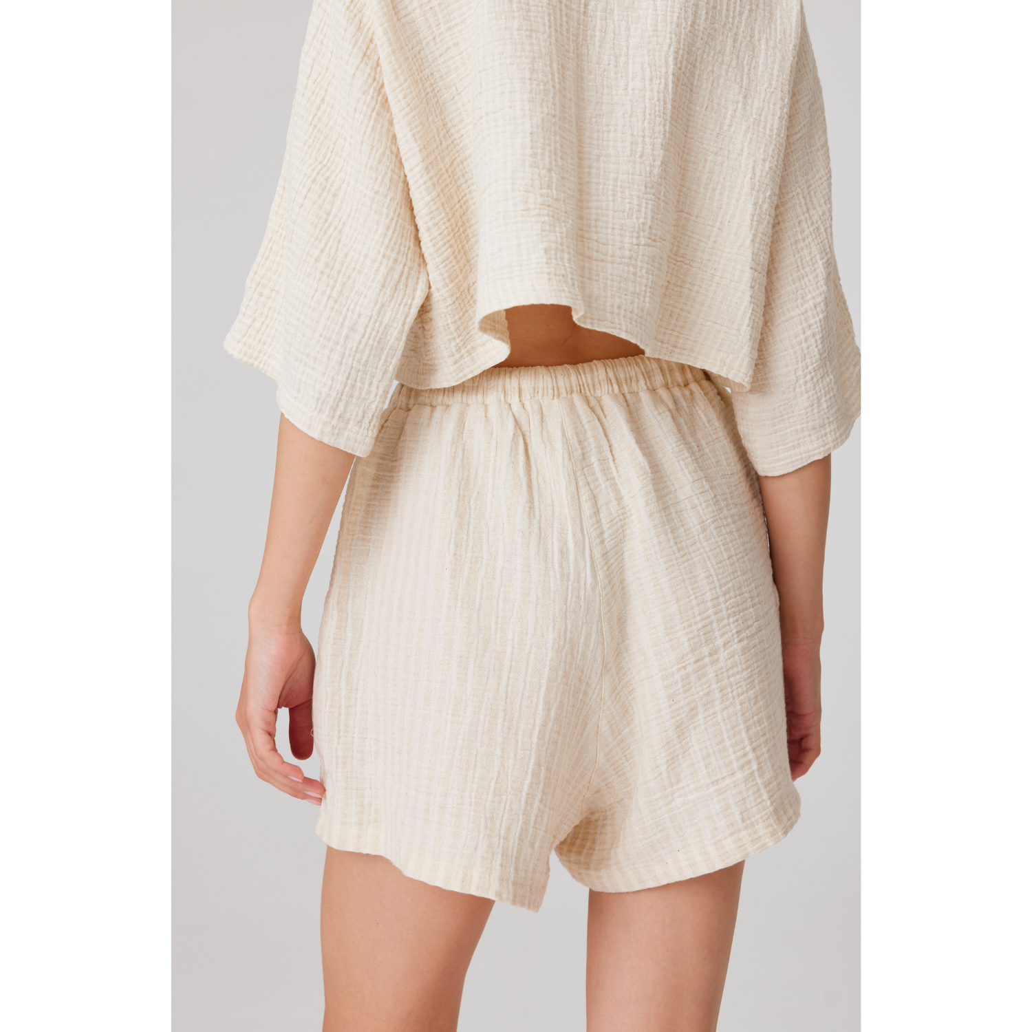 Rear view of a woman wearing a cream-colored cotton linen shorts set, showcasing the relaxed fit of the high-waisted shorts and the loose drape of the cropped button-up summer shirt.