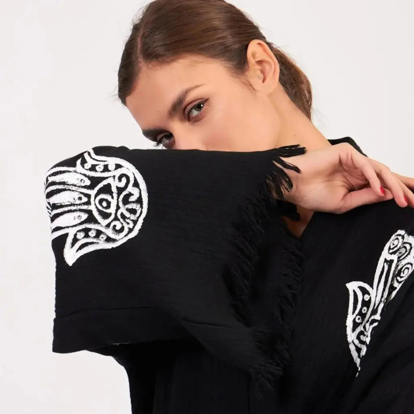 Une jeune femme portant un kimono Trimita noir et à motifs avec un motif Hamsa blanc (Main de Fatma) et des détails de franges.Livraison gratuite dès 75€ d'achat.