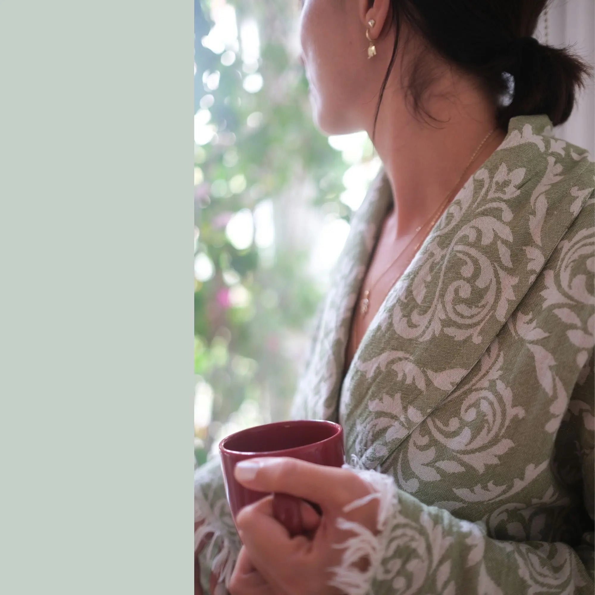 A woman standing by a bright window in a light green baroque patterned bathrobe, holding a red mug, embodying a luxury home spa and relaxation vibe.