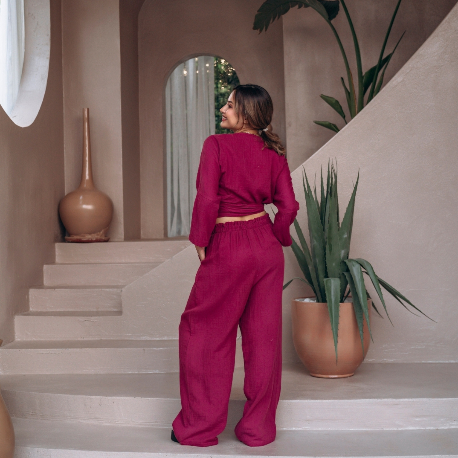 Rear full-length view of a woman in a premium magenta pink muslin co-ord set, displaying the smooth flowy fit of the wide-leg pants and the back layout of the long-sleeve crop top.