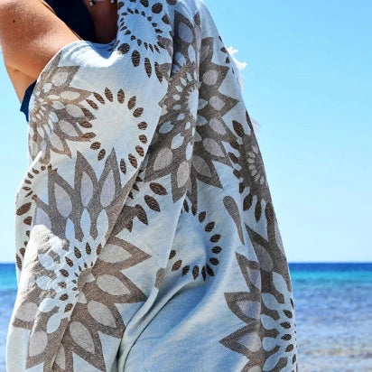 A close-up shot of a person wrapped in the light blue sunflower-patterned Turkish towel (Pestemal) at the beach, with the turquoise sea and blue sky in the background.