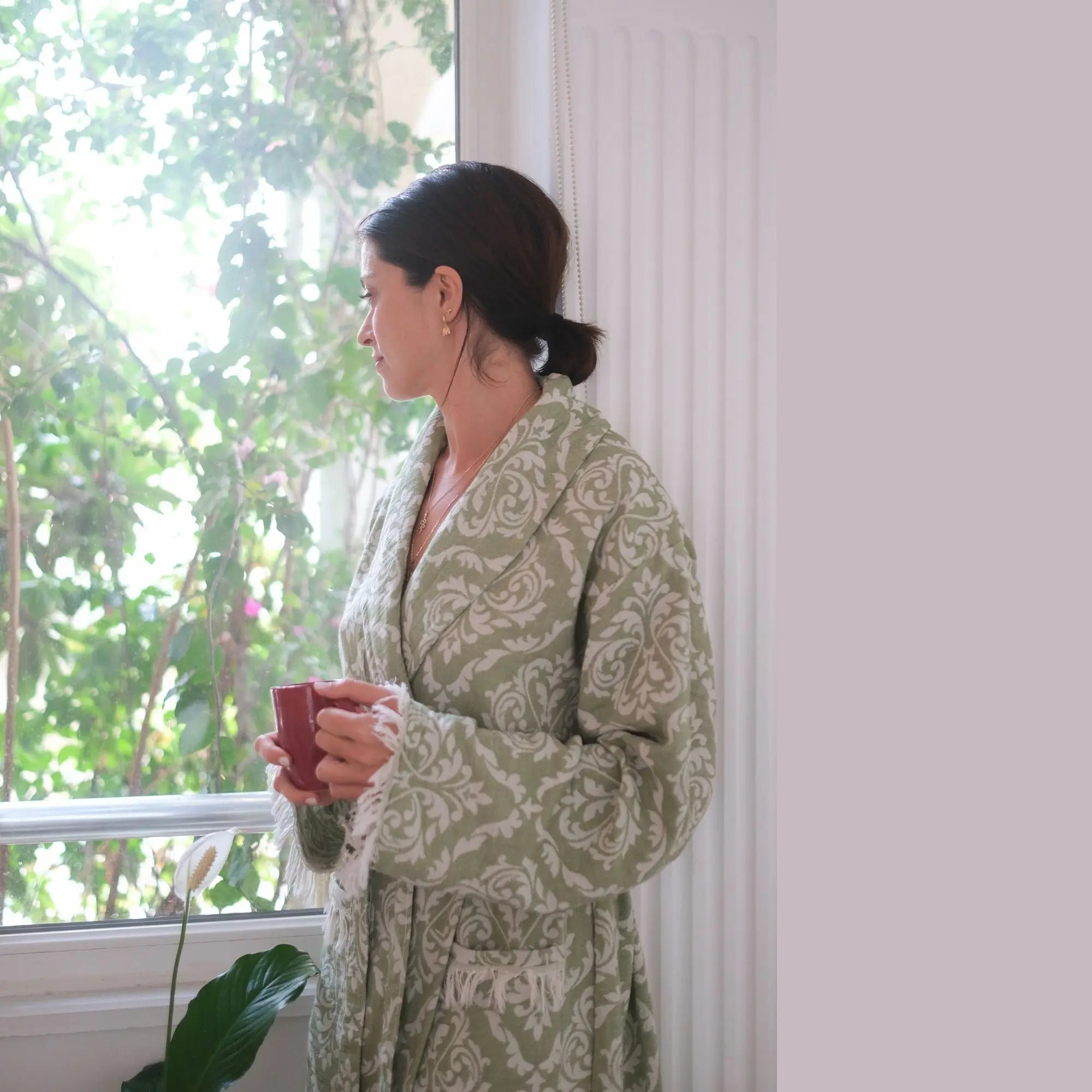 Close-up of a person wearing a sage green Turkish peshtemal robe while holding a coffee mug, focusing on the belted waist and textured pocket detail.