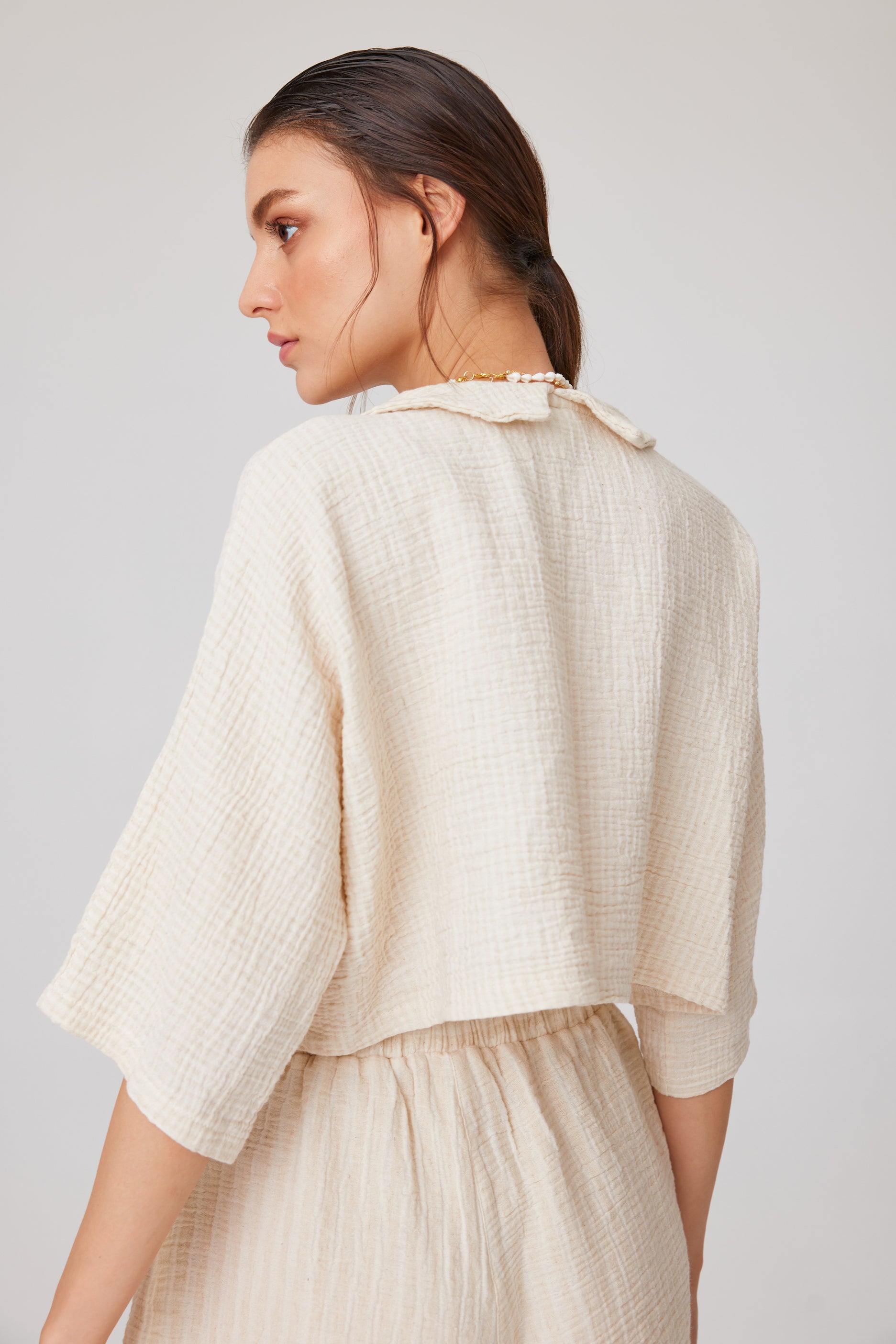 Person with long dark hair styled back, wearing a neutral-toned, loose-fitting short set with button-up top and drawstring shorts in a minimalist studio setting