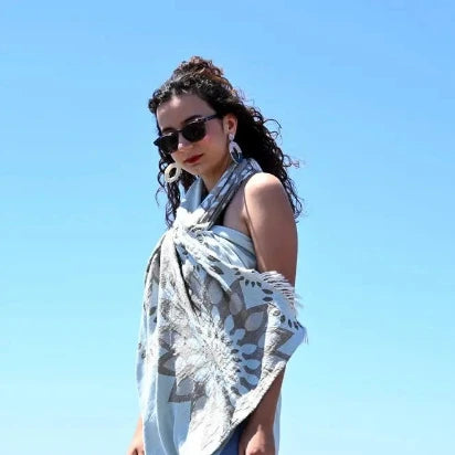 A stylish woman with dark curly hair wears the light blue sunflower Turkish towel wrapped around her body like a dress, accessorized with sunglasses and large white earrings, against a clear blue sky.