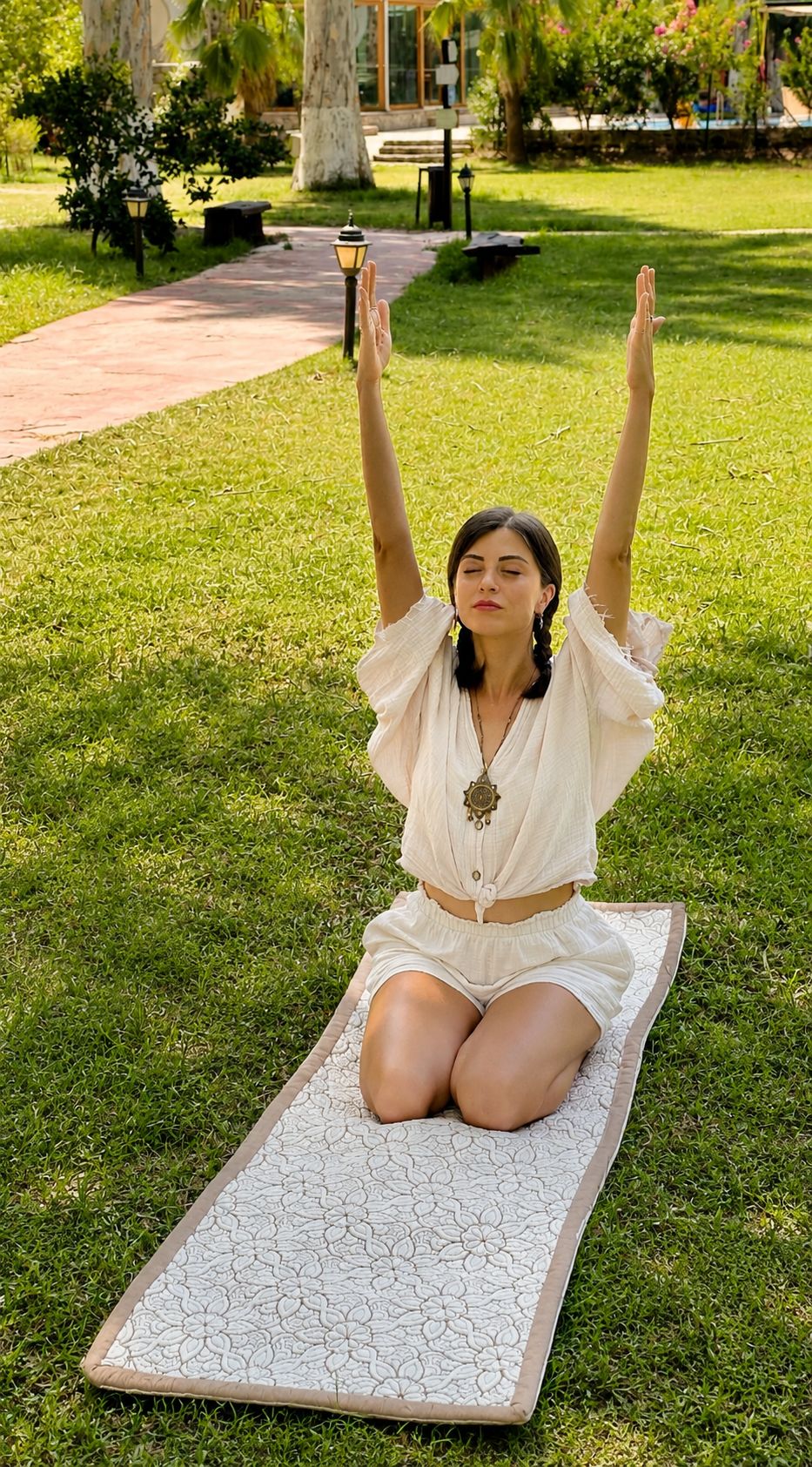 Vrouw knielt met geheven armen op een crèmekleurige Lotus yogamat met bloemenpatroon op het gras.
