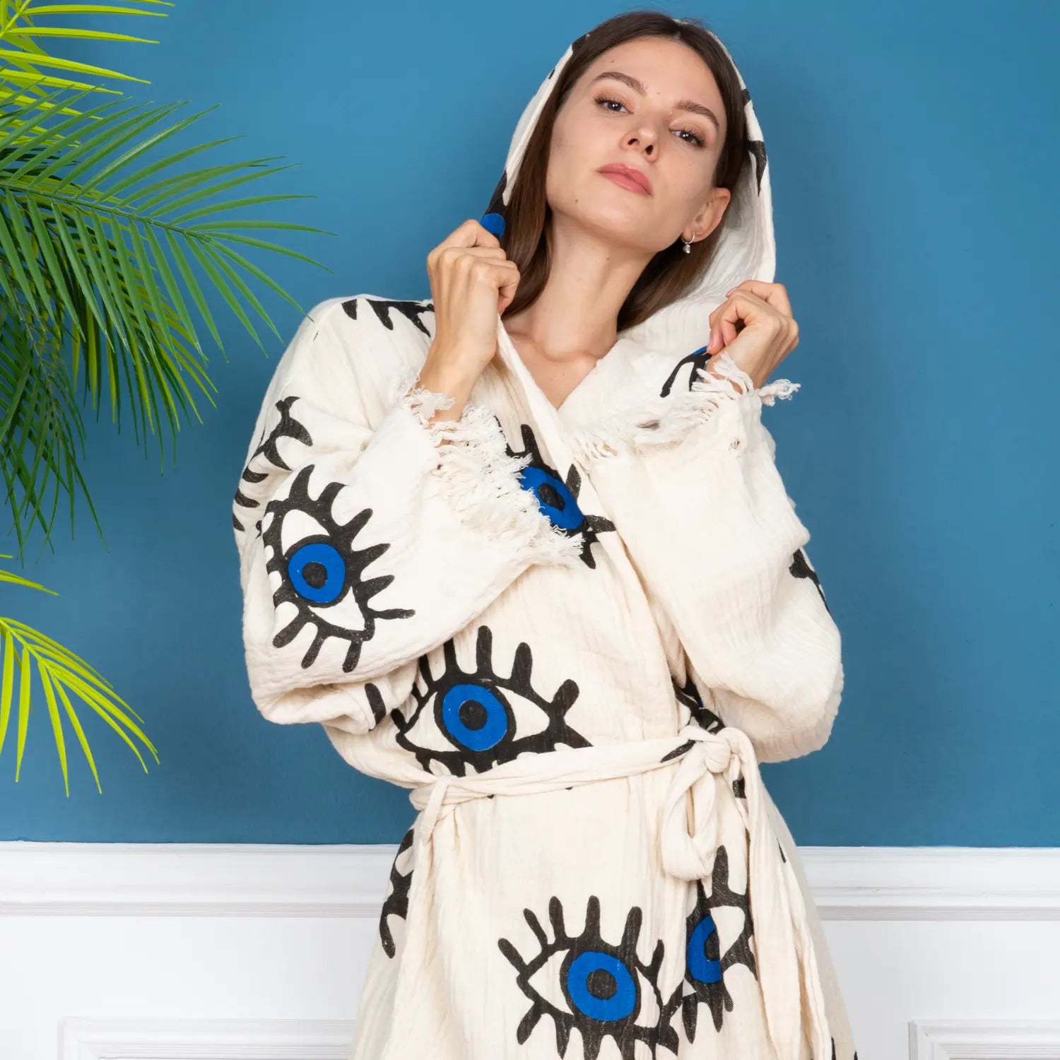 A young woman modeling a creamy white muslin hooded bathrobe with all-over blue and black evil eye pattern, belted at the waist, posing against a blue wall with a palm leaf in the background.Free shipping on orders over $100.