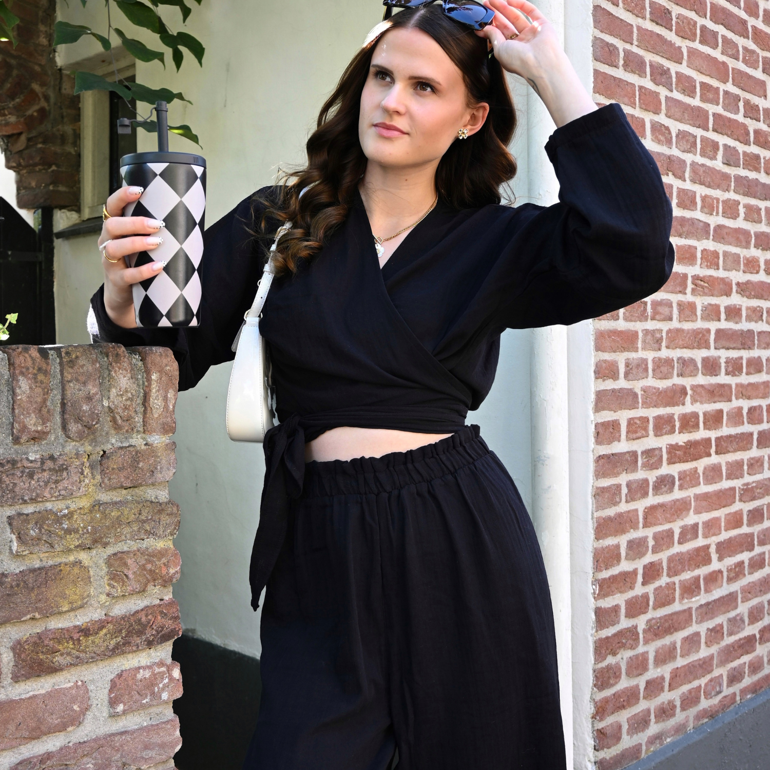 Trendy woman outdoors wearing a black breathable cotton muslin crop top and high-waisted relaxed trousers, holding a coffee mug and touching her sunglasses.