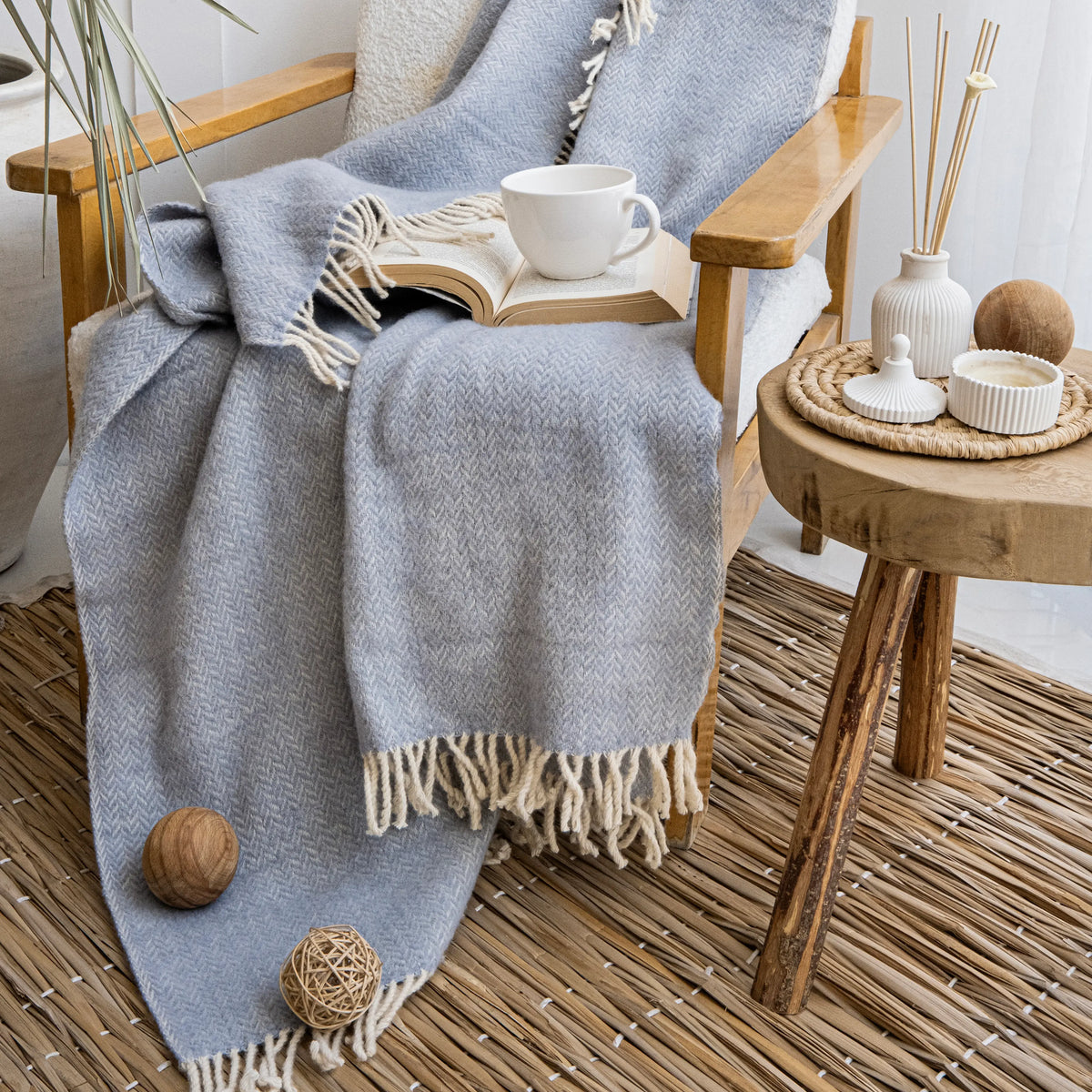 Light-blue Ladira wool throw draped over a wooden chair beside a book and a cup.
