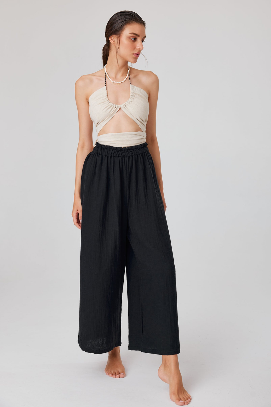Femme portant un haut clair à encolure dos-nu et découpe, avec un pantalon noir à jambes larges sur fond uni.