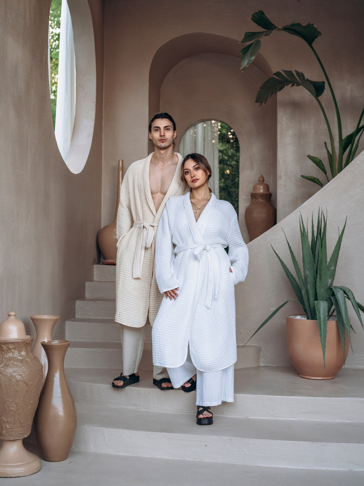 Two people in Trimita Soft Waffle bathrobes standing on a staircase with decorative vases and plants.