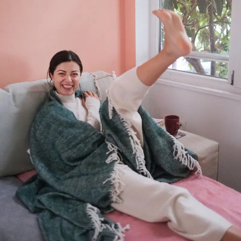 Sevgin zit op de bank met een groene deken van Trimita, lacht en strekt haar been uit.