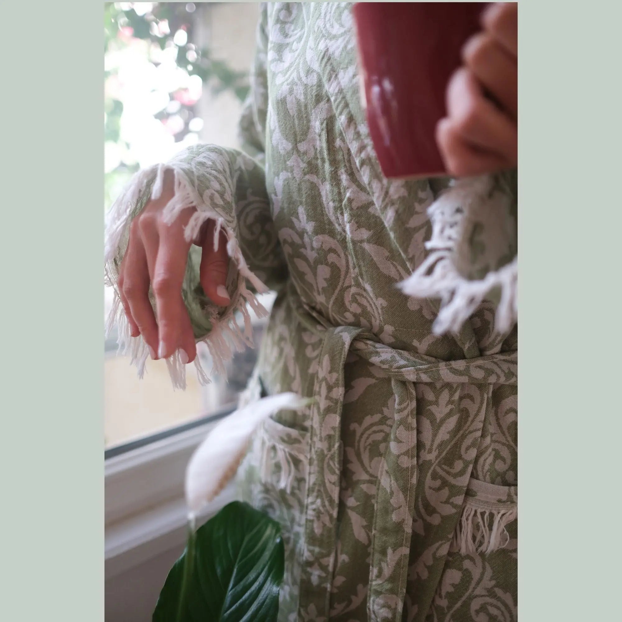 A woman standing by a bright window in a light green baroque patterned bathrobe, holding a red mug, embodying a luxury home spa and relaxation vibe.