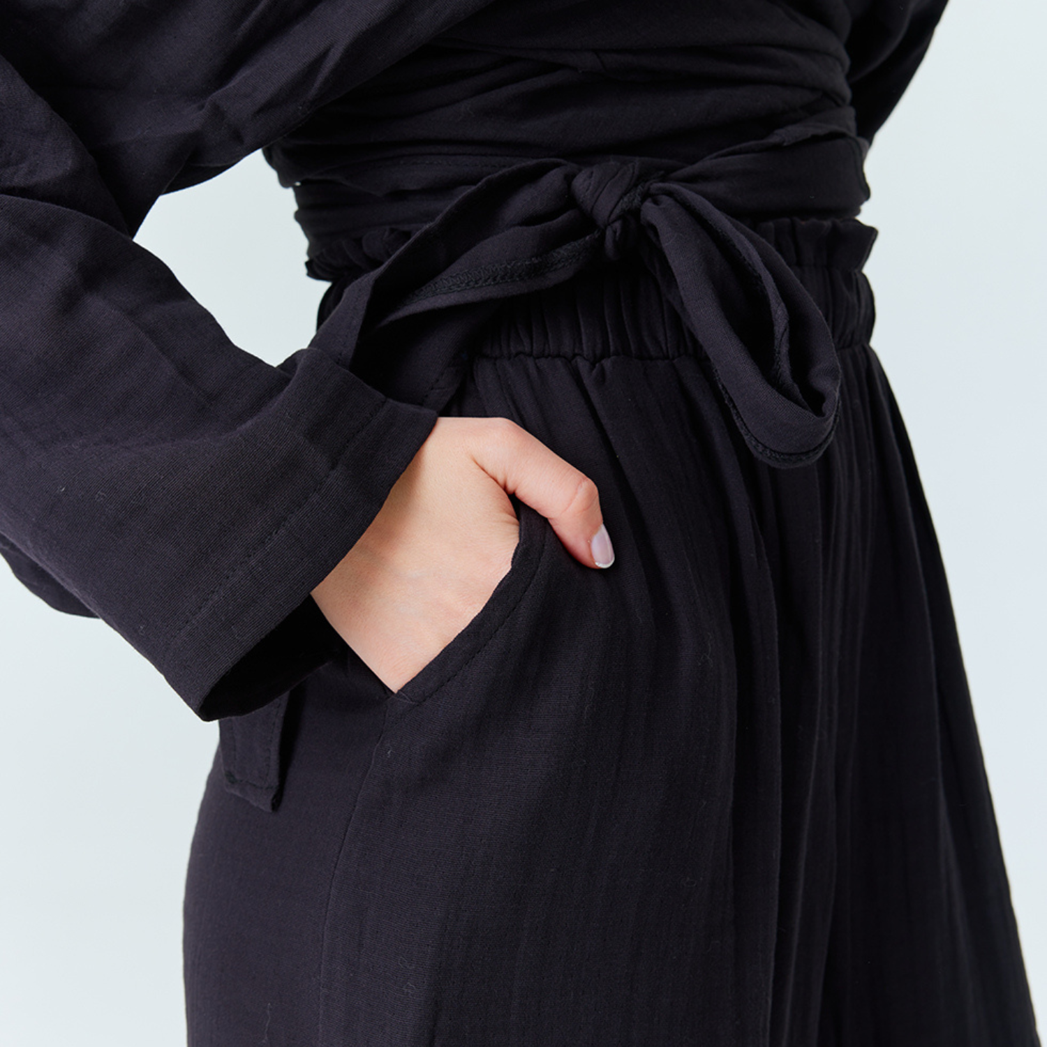 Close-up structural view highlighting the functional side pocket, flexible elastic waistband, and tie-knot bow of the black cotton muslin loose-fit pants.