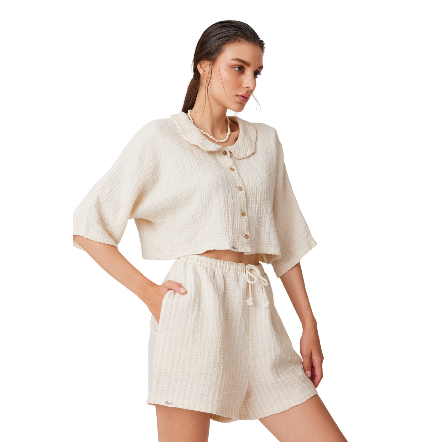 Full-body studio shot of a model posing in the Countryside Breeze cream cotton linen shorts set, featuring an open-collar cropped shirt with wooden buttons and matching high-waisted relaxed shorts.