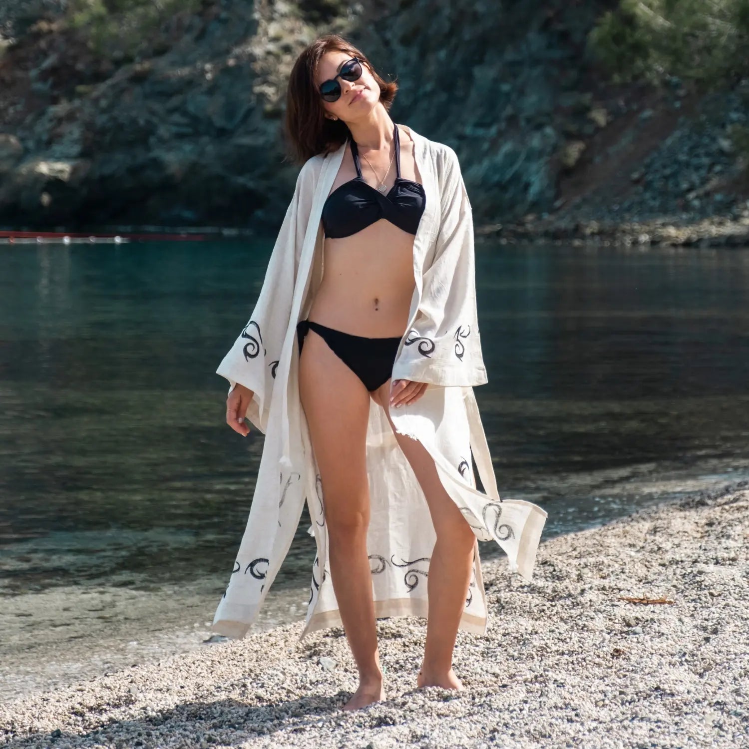 Frau in einem schwarzen Bikini und Sonnenbrille, die einen leichten, cremefarbenen Strandkimono mit schwarzen Wirbelmustern trägt, stehend an einer felsigen Bucht mit türkisfarbenem Wasser. Kostenloser Versand bei Bestellungen über 50€.