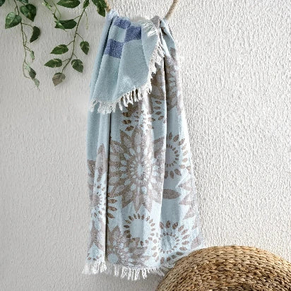 The light blue Turkish towel (Pestemal) with the brown sunflower pattern and blue stripes hangs on a rustic rope hook against a textured white wall, with green leaves on a vine to the side.