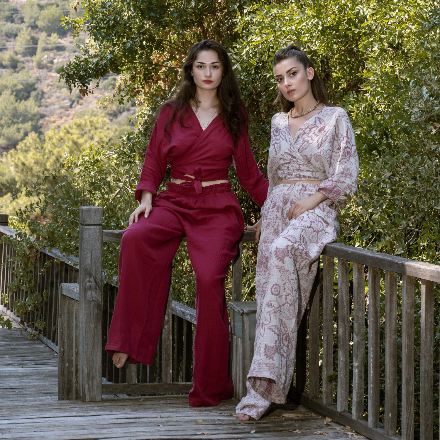 Two women posing on a wooden bridge; one wearing a solid magenta cotton muslin wrap crop top with wide-leg pants, and the other wearing a patterned beige and rose muslin coordinated set.