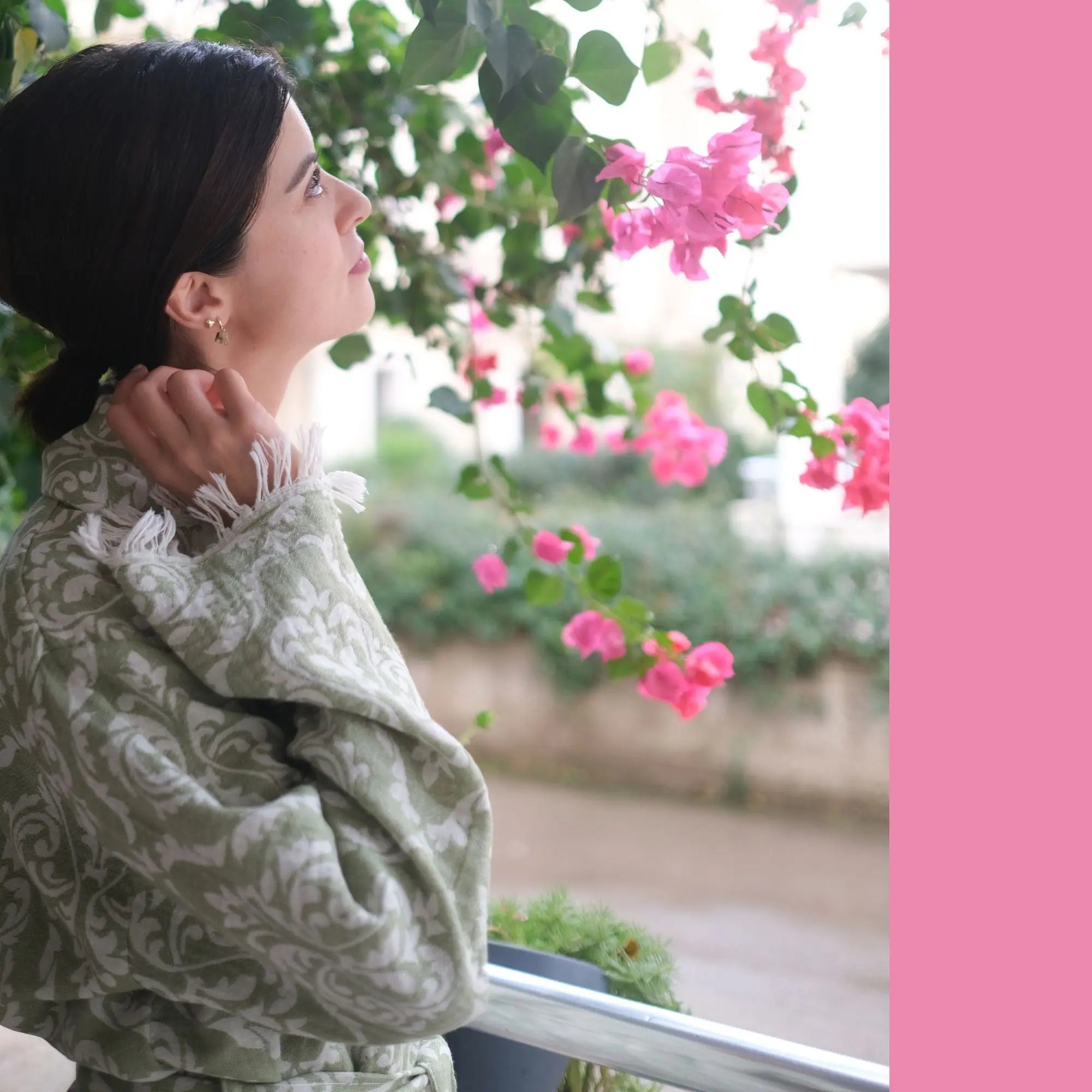 A woman standing on a balcony wearing a green handmade Turkish cotton bathrobe, looking at pink bougainvillea flowers in a serene morning setting.